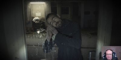 Film still from 'First Person' (2023) by Jon Beaton. A bearded man with closed eyes is seen outside a window at night. The man is gesturing that he is going to bed by holding his hands together against his cheek.